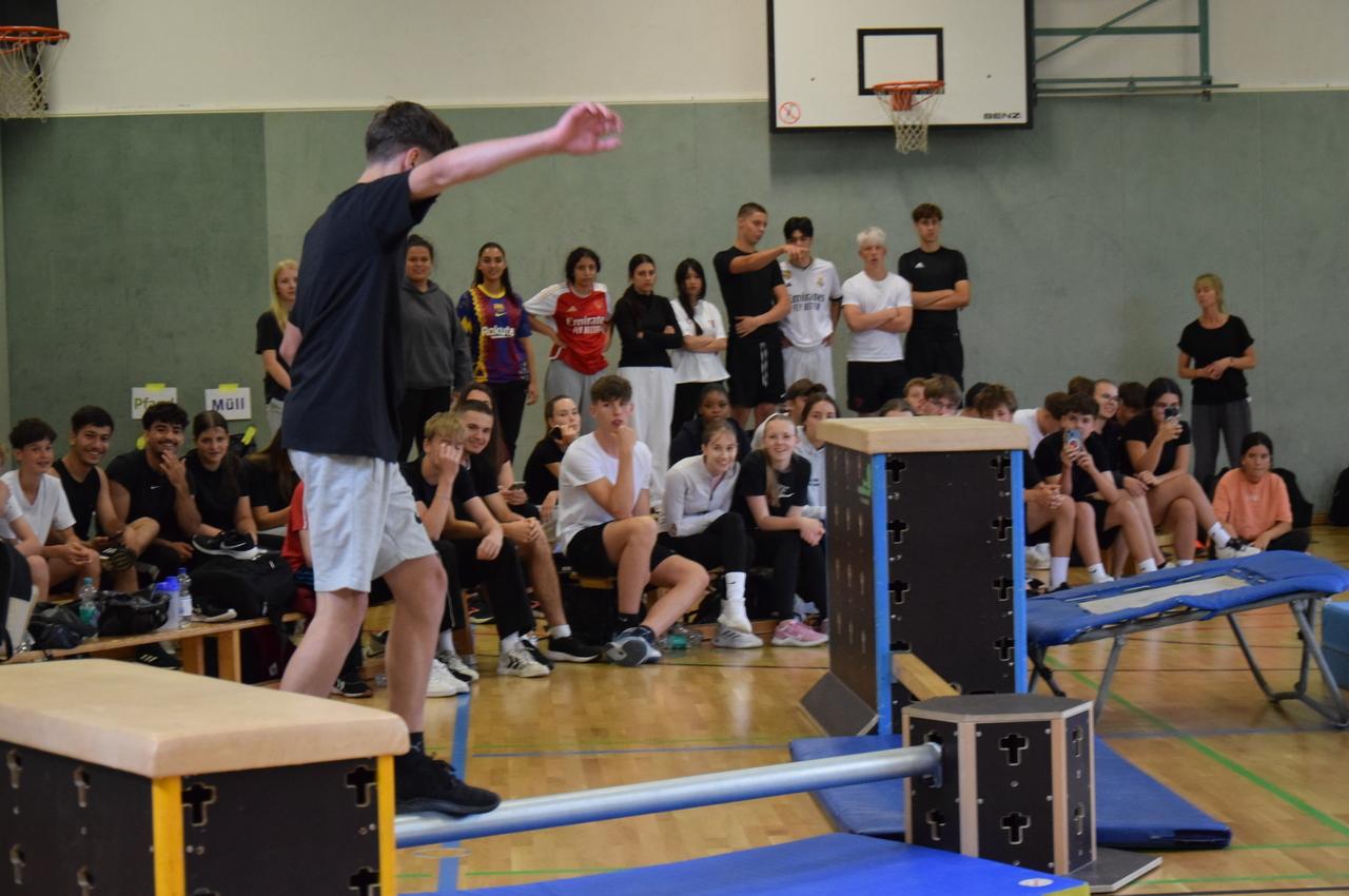 Junge balanciert auf einem schmalen Balken, umgeben von zuschauenden Schülern in einer Sporthalle.