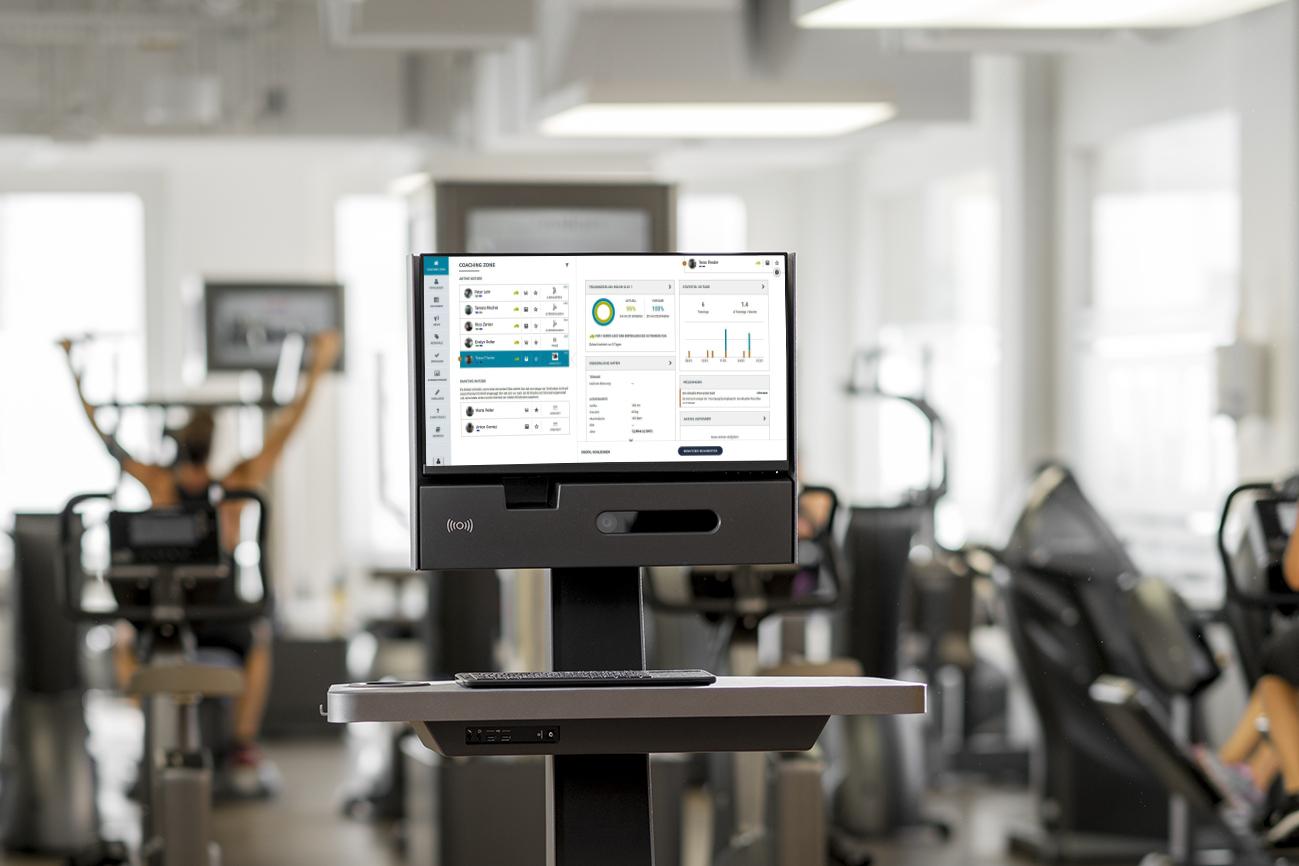 Fitnessstudio mit einem monitorisierten Fitnessgerät auf einem stabilen Tisch. Im Hintergrund trainierende Personen.