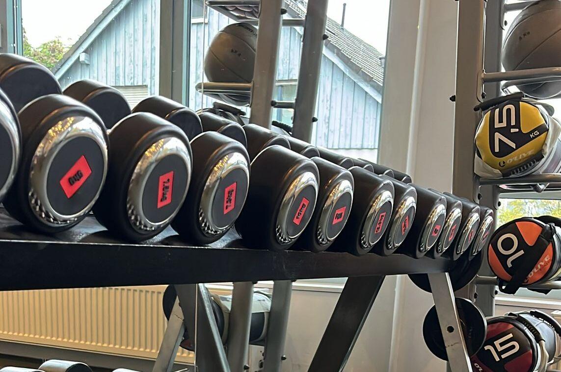Reihe von Kurzhanteln mit schwarzen Griffen auf einem Rack in einem Fitnessstudio, Spiegel an der Wand im Hintergrund.