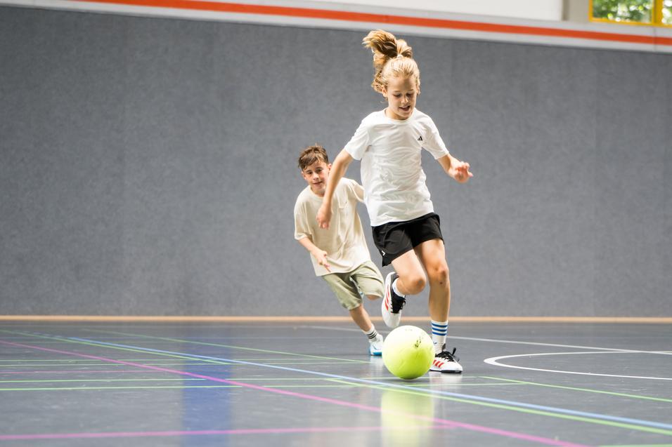 Zwei Kinder spielen in einer Halle Fußball, ein Kind rennt dem Ball hinterher, während das andere zuschaut.