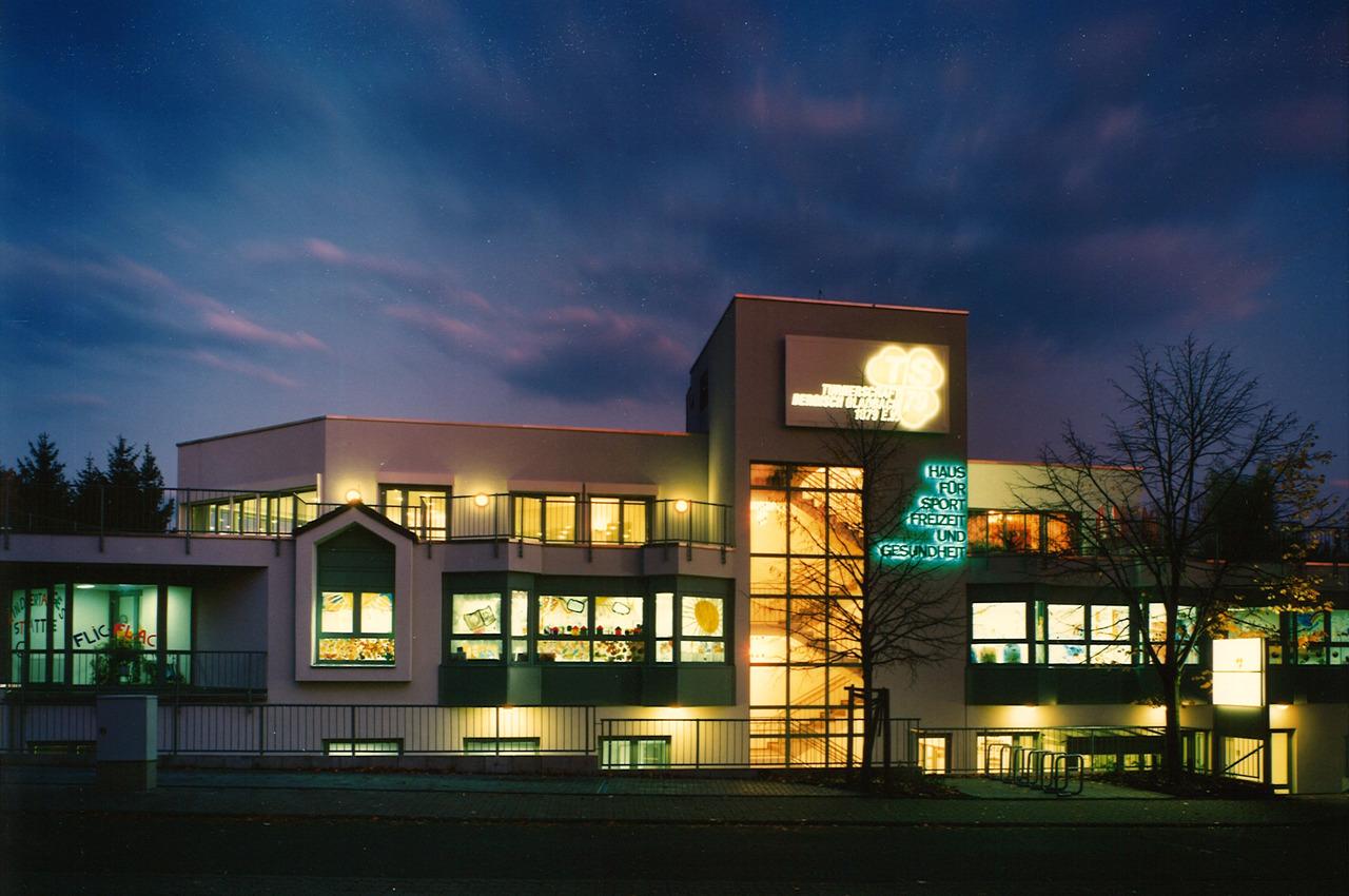 Moderne Sporteinrichtung bei Dämmerung, mit beleuchteten Fenstern und dem Schriftzug „Haus für Sport, Freizeit und Gesundheit“.