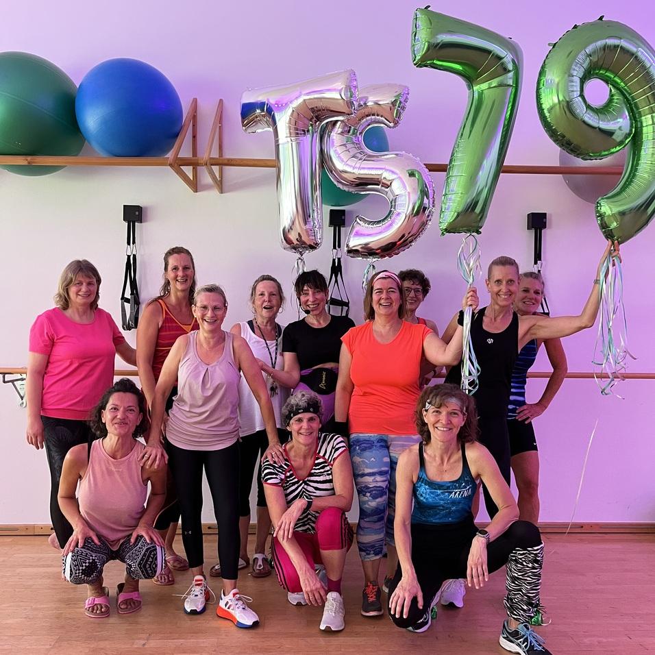 Gruppe von 12 Frauen in Sportkleidung posiert vor bunten Luftballons mit der Aufschrift „TS 79“ in einem Fitnessraum.